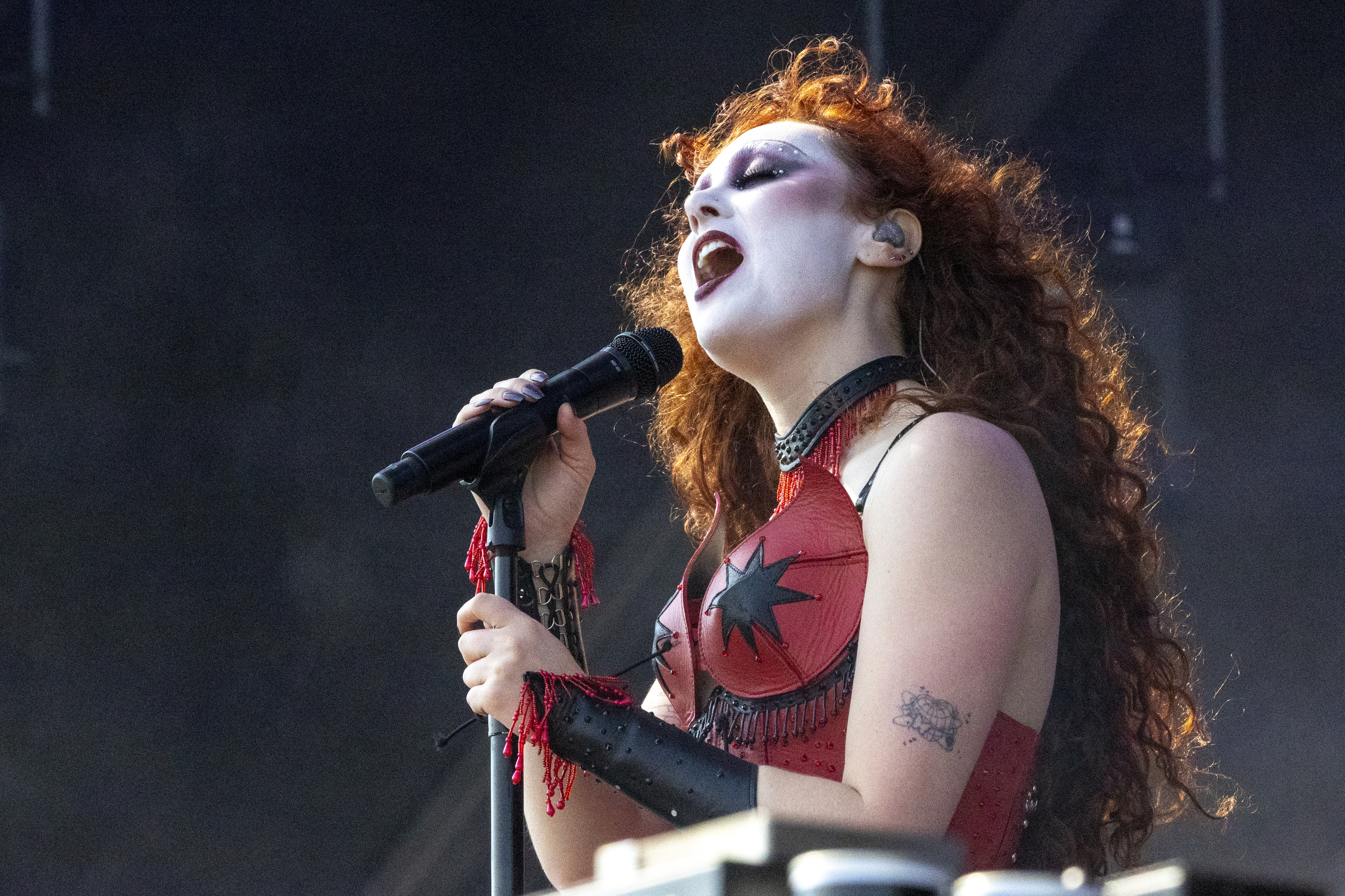 Chappel Roan performs on stage, passionately singing into a microphone. She is dressed in a dramatic red outfit with a star design, showcasing bold makeup and wavy hair