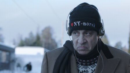 Steven Van Zandt, wearing a "I ♥ NY-norsk" beanie and headphones, stands outside in a winter setting, bundled in a jacket and scarf