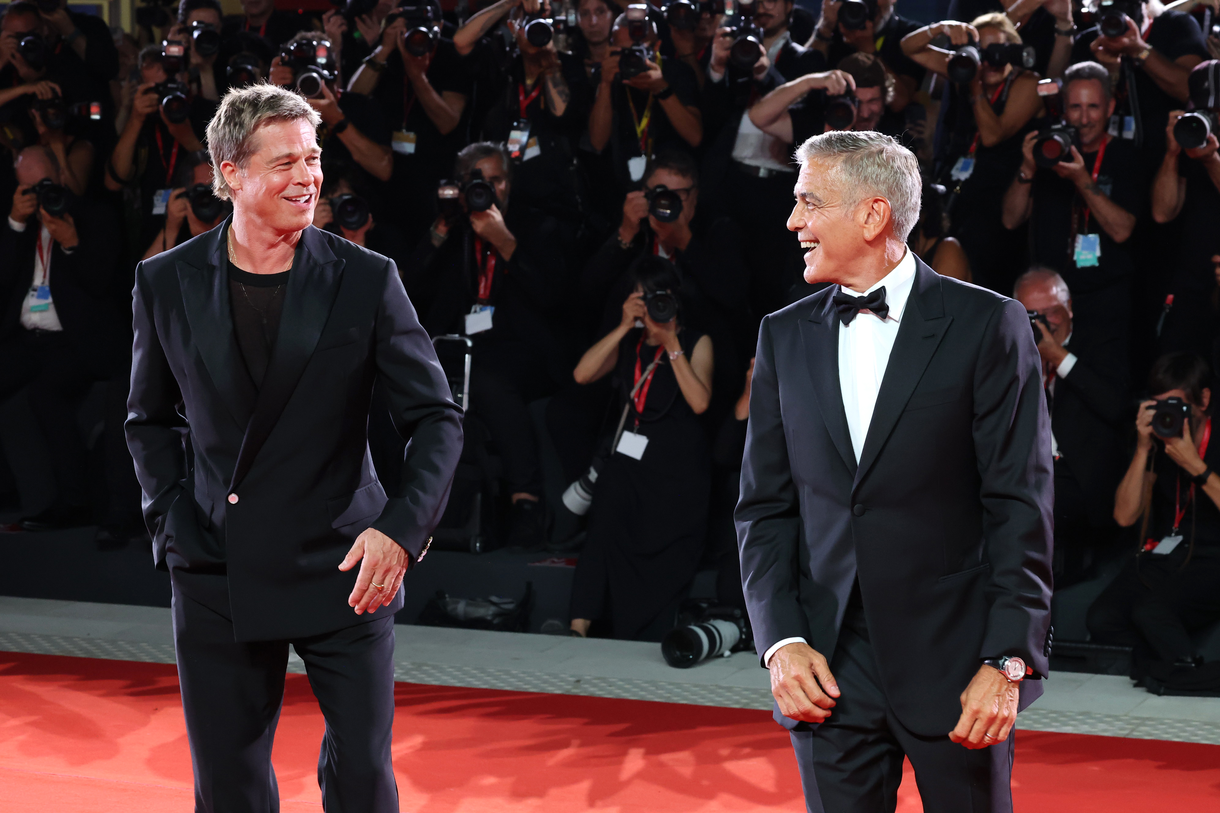 Brad Pitt and George Clooney smiling on the red carpet with photographers in the background