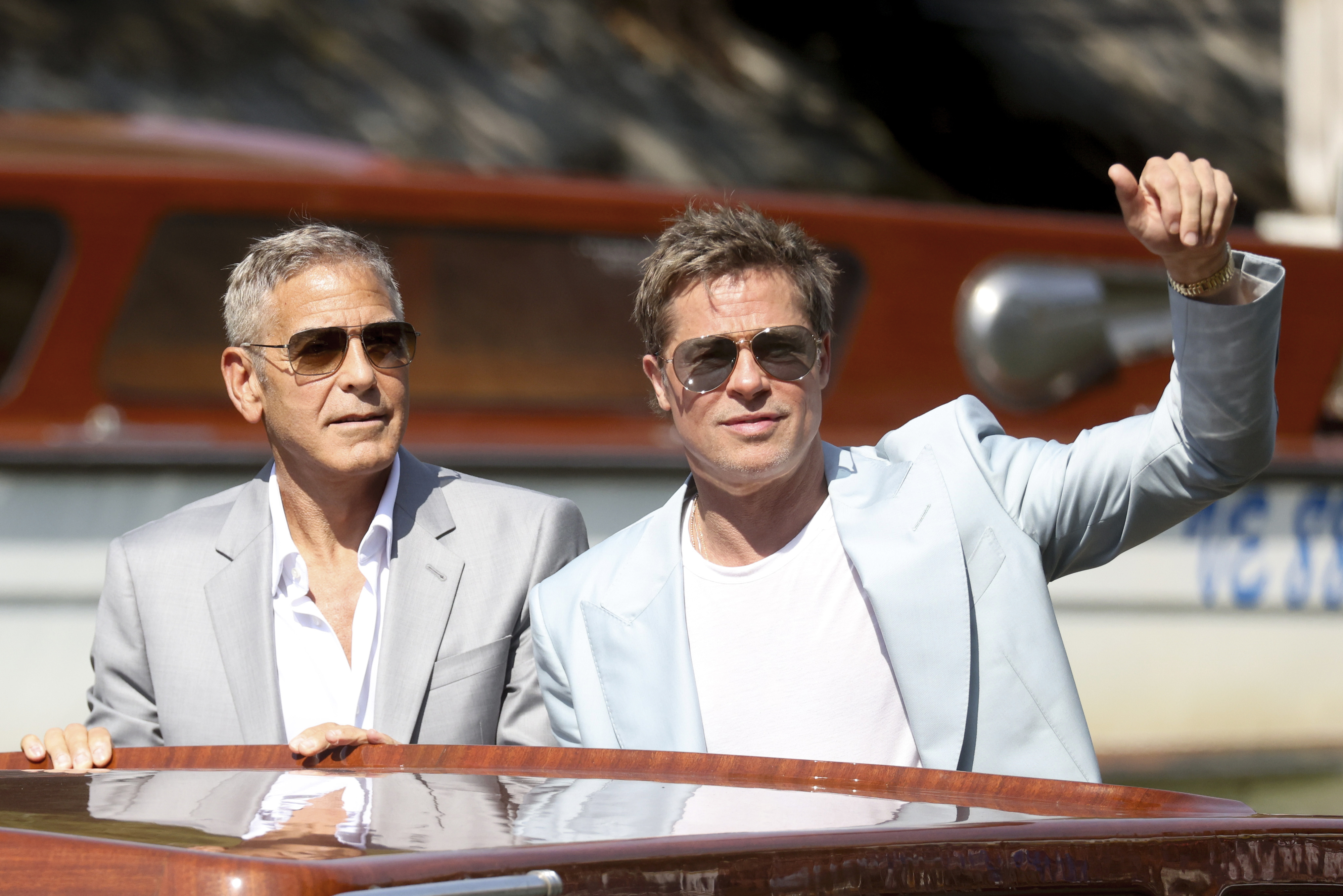 George Clooney and Brad Pitt on a boat; Clooney in a light suit and sunglasses, Pitt in a light jacket and sunglasses, waving
