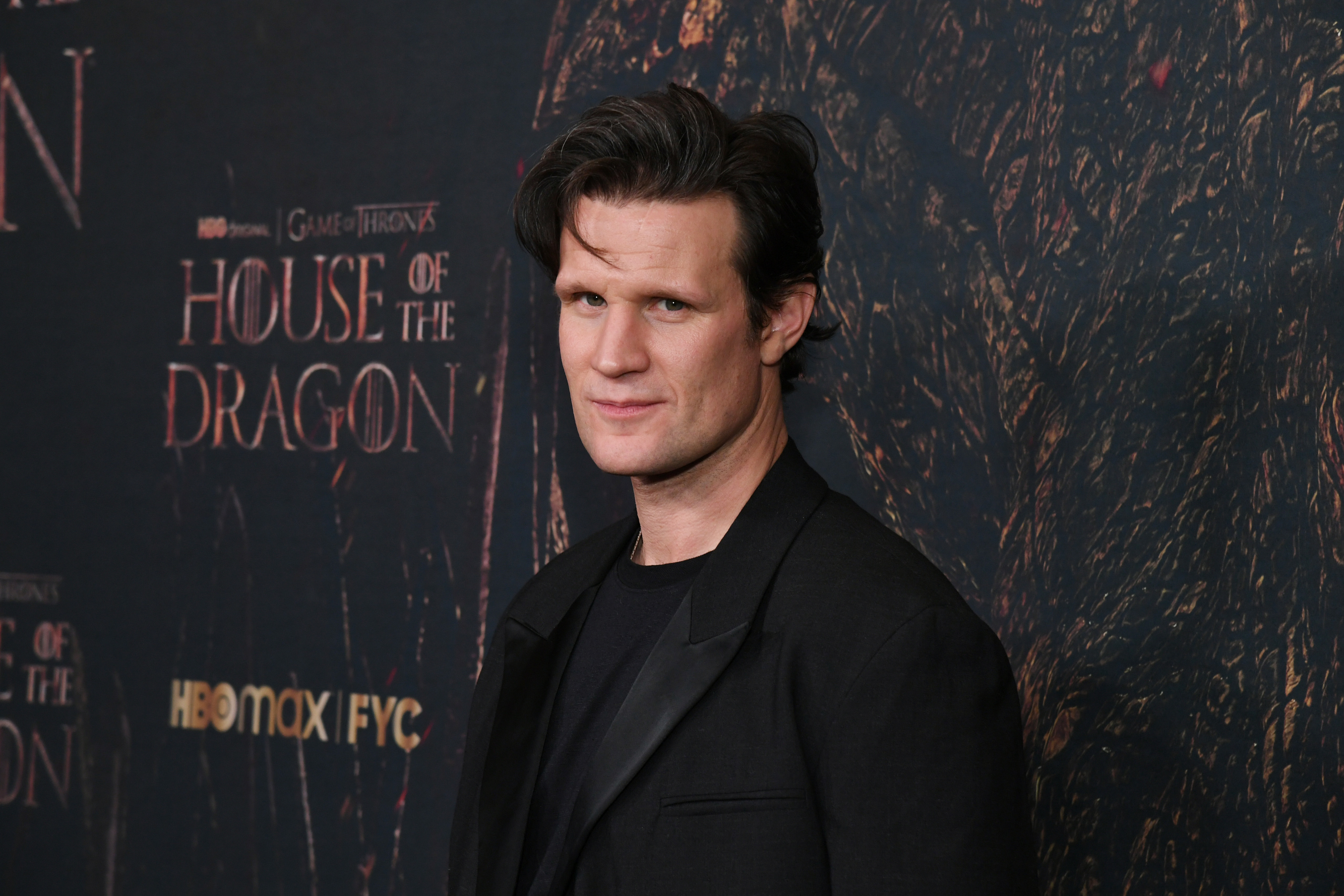 Matt Smith at the &quot;House of the Dragon&quot; premiere event, wearing a black suit jacket over a black shirt, standing in front of an event backdrop