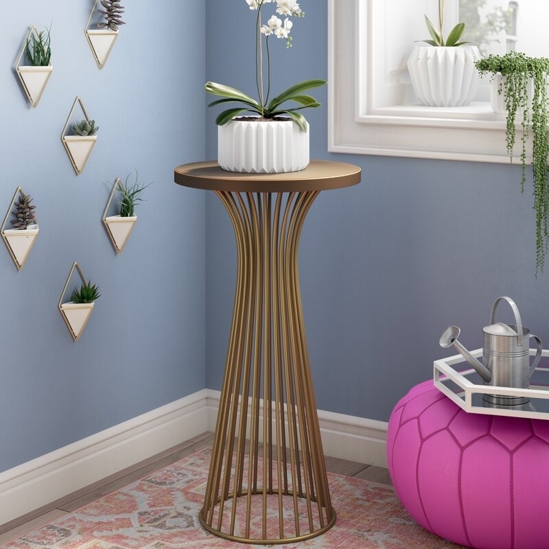 A modern side table with a potted orchid on top stands beside a window with hanging plants. There are small wall planters and a pink pouf nearby