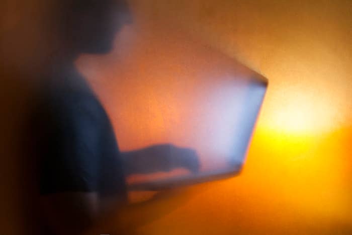 A person uses a laptop, appearing blurred and silhouetted through a frosted or smoky screen. Faces and detailed features are not visible