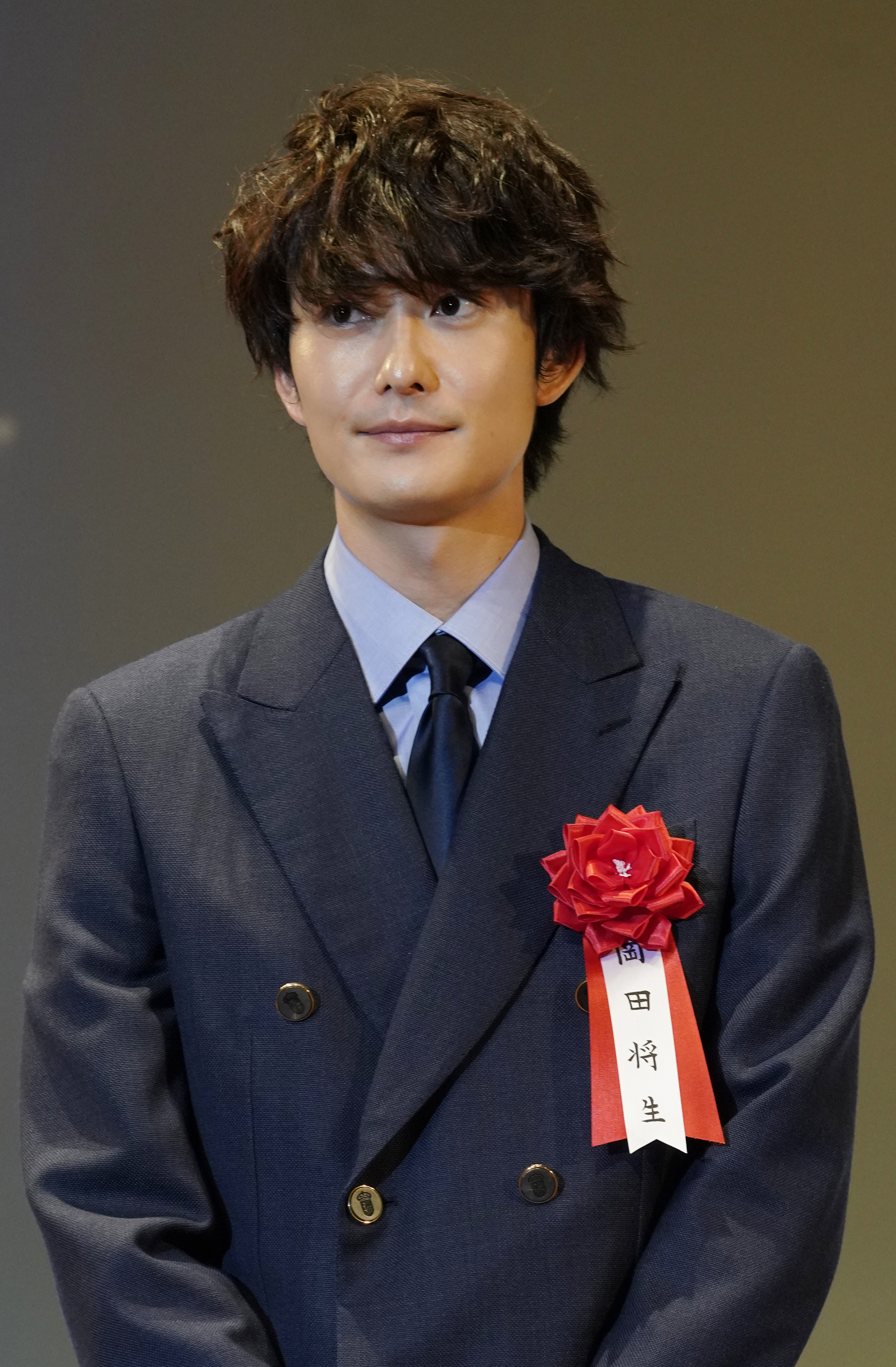 向井理がダブルジャケットのスーツを着用し、胸に赤いリボン付きの名札をつけている。