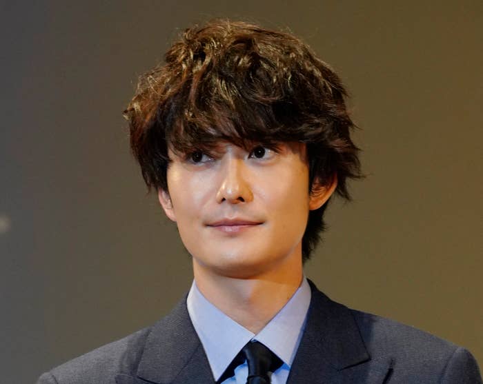 向井理がダブルジャケットのスーツを着用し、胸に赤いリボン付きの名札をつけている。