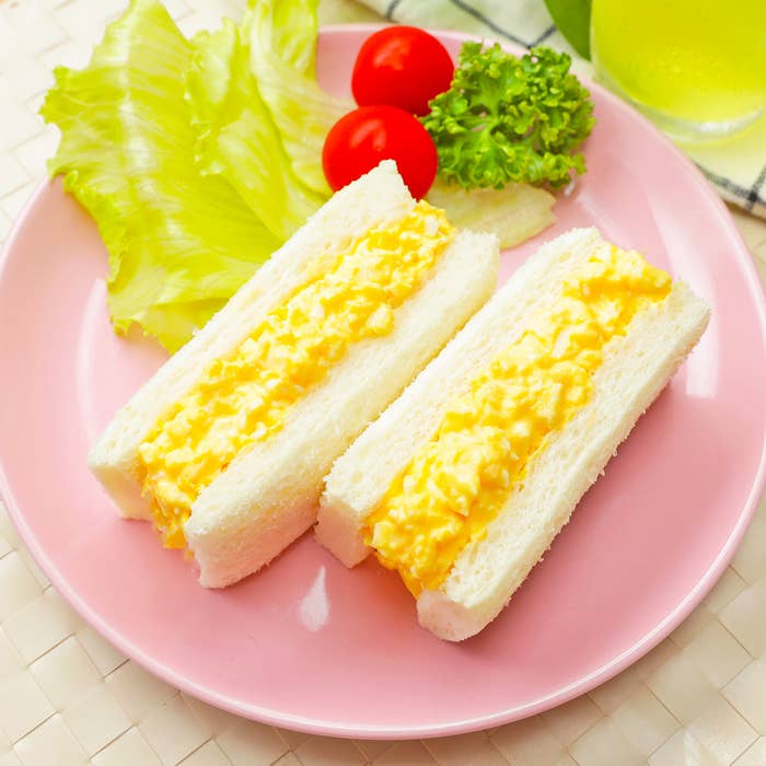 ピンクの皿にのった卵サンドイッチと、レタス、プチトマト、パセリの付け合わせ。