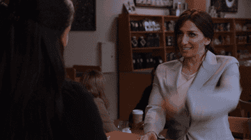 Two women in a coffee shop: one in a white blazer enthusiastically gesturing with her arms, another facing away with long hair