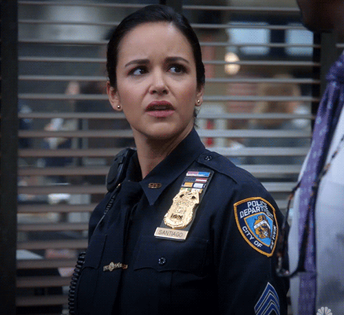 Stephanie Beatriz as Amy Santiago in a police uniform, looking puzzled on the set of Brooklyn 99, with visible badge and patches