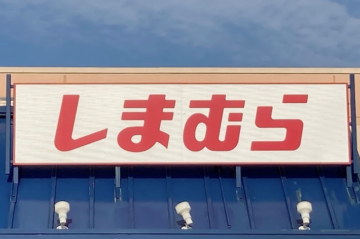 Storefront with a sign displaying Japanese text. Large red &ldquo;しまむら&rdquo; characters are on a white background. Building has accessible parking
