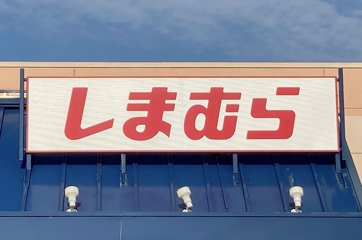 A storefront sign with red Japanese characters reads &ldquo;しまむら.&rdquo; The building features blue awnings and a handicapped parking sign