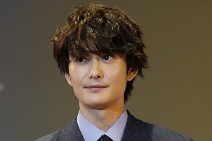 向井理がダブルジャケットのスーツを着用し、胸に赤いリボン付きの名札をつけている。