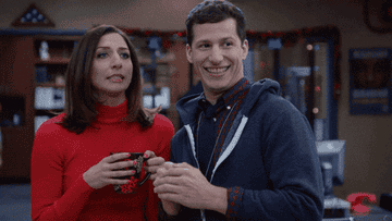 Chelsea Peretti holds a mug while standing next to Andy Samberg, who is smiling, in a festive indoor setting