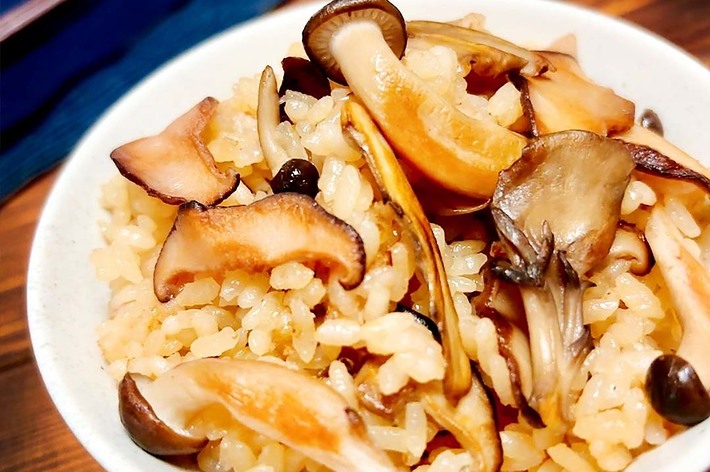 きのこご飯の近接画像。白いボウルに盛られた炊き込みご飯に、数種類のきのこが添えられている。背景に箸と布が見える。