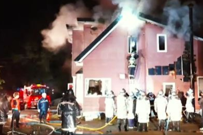 消防隊員が夜間に火事のあった家屋で消火活動中。家屋の2階から煙が立ち上り、多くの消防士がホースを使い対応している。
