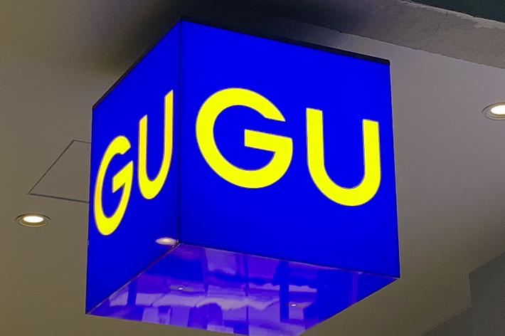 Square blue sign with yellow &ldquo;GU&rdquo; letters, seen hanging from a ceiling