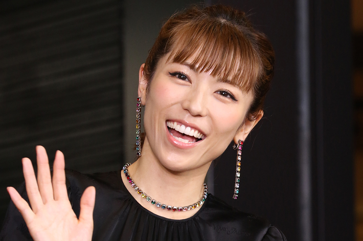 女性が赤い花の飾りをつけた黒いドレスを着て笑顔で両手を振る姿。