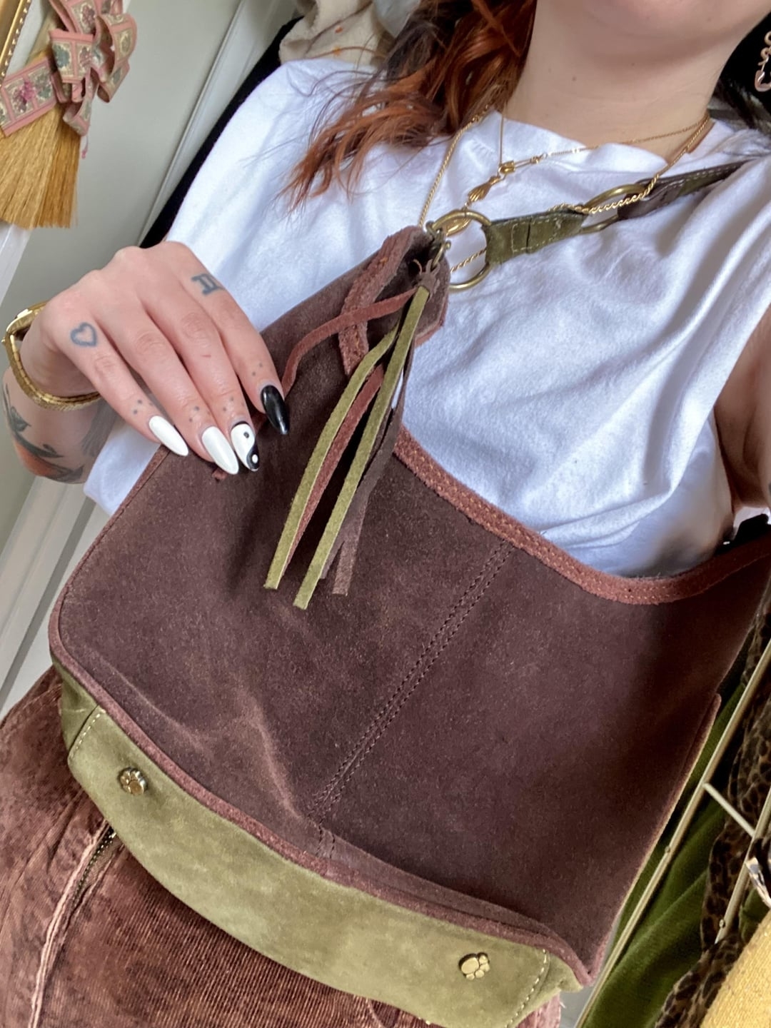 A person with nail art and tattoos is holding an earthy-toned suede bag