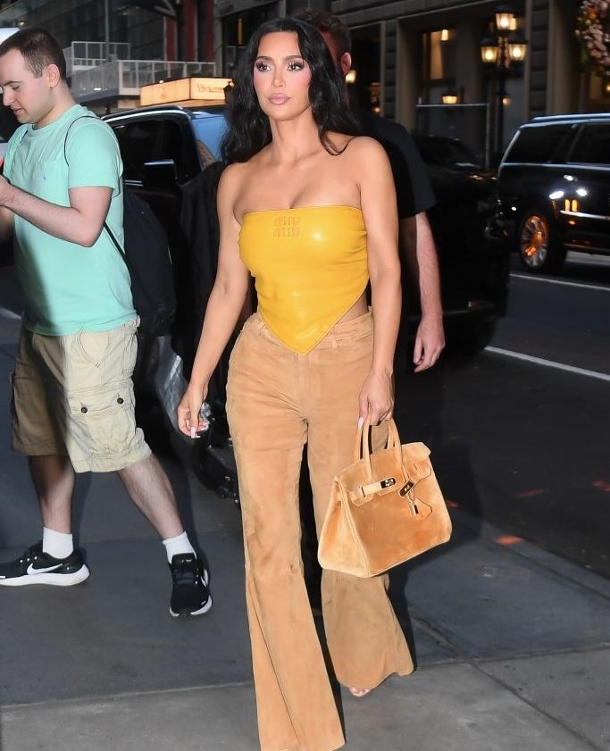 Kim Kardashian walks on a city street wearing a strapless yellow top, brown pants, and holding a large tan handbag