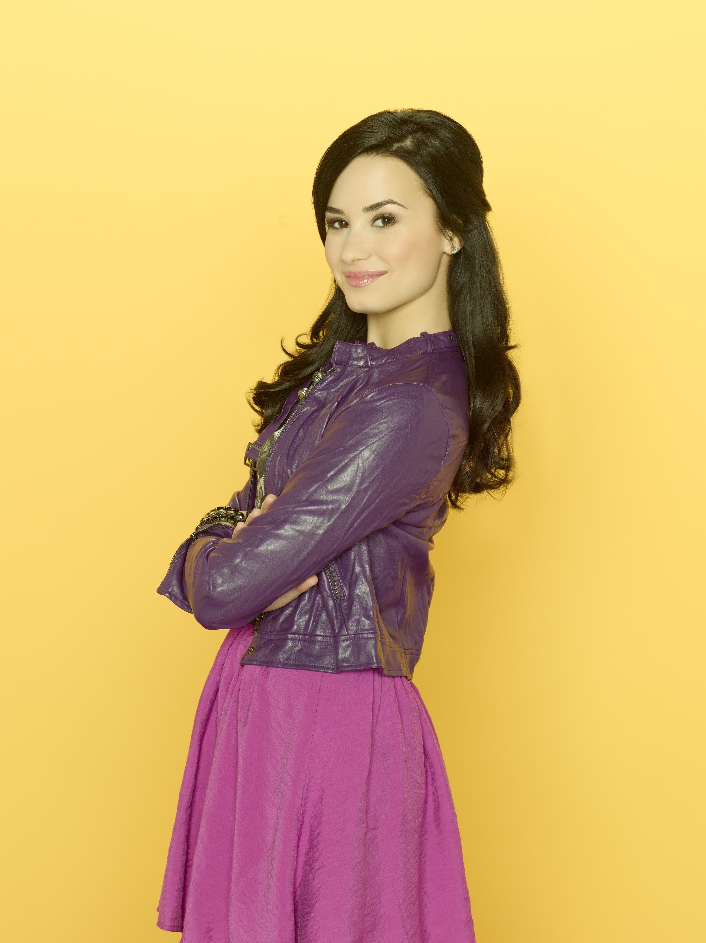 Demi Lovato smiling with arms crossed, wearing a leather jacket and a dress against a plain background