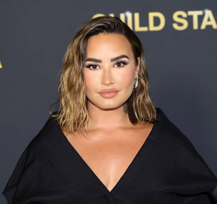 Demi Lovato stands in front of a "Child Star" sign, wearing an elegant black off-shoulder gown, with waved hair, neutral makeup, and statement earrings