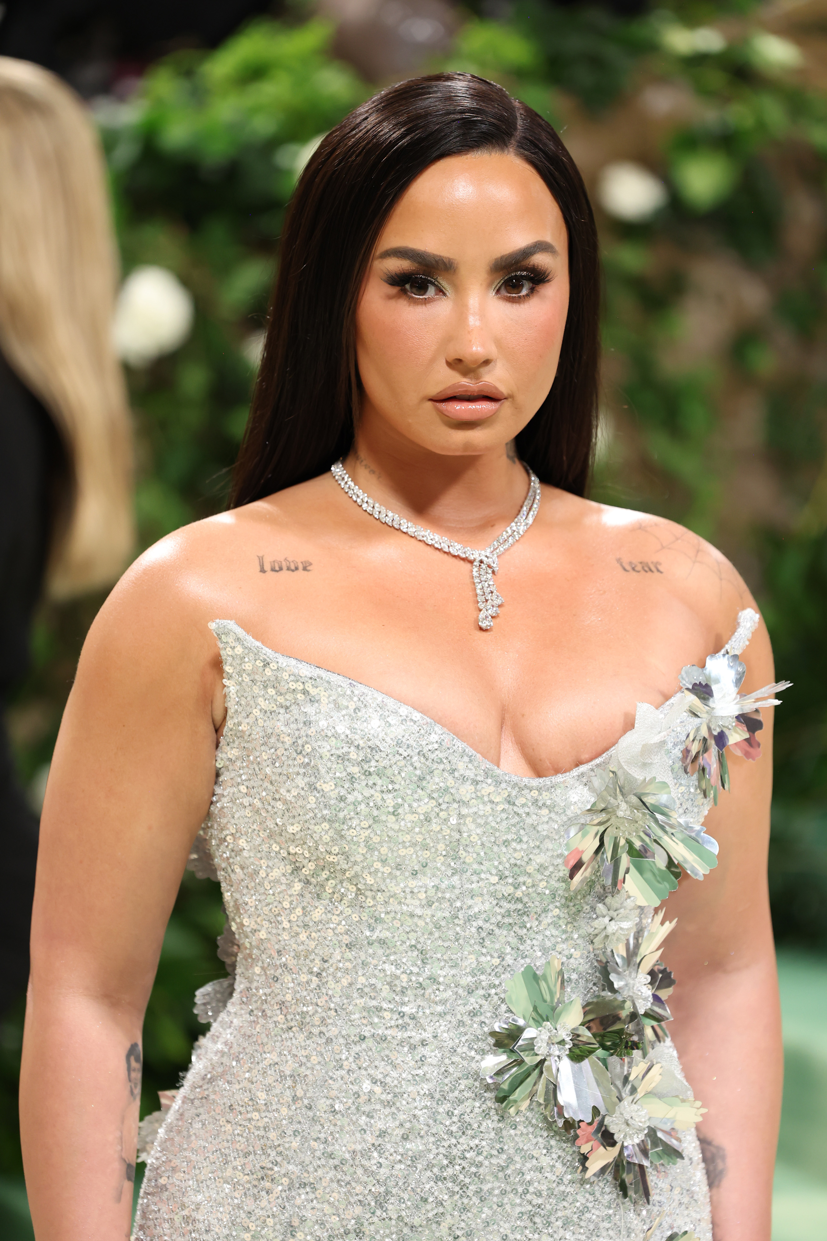 Demi Lovato on the red carpet, wearing a strapless sparkling gown adorned with floral appliqués and a diamond necklace