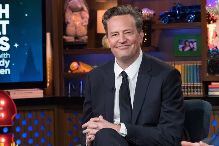 Matthew Perry, in a suit and tie, smiles while seated on a talk show set
