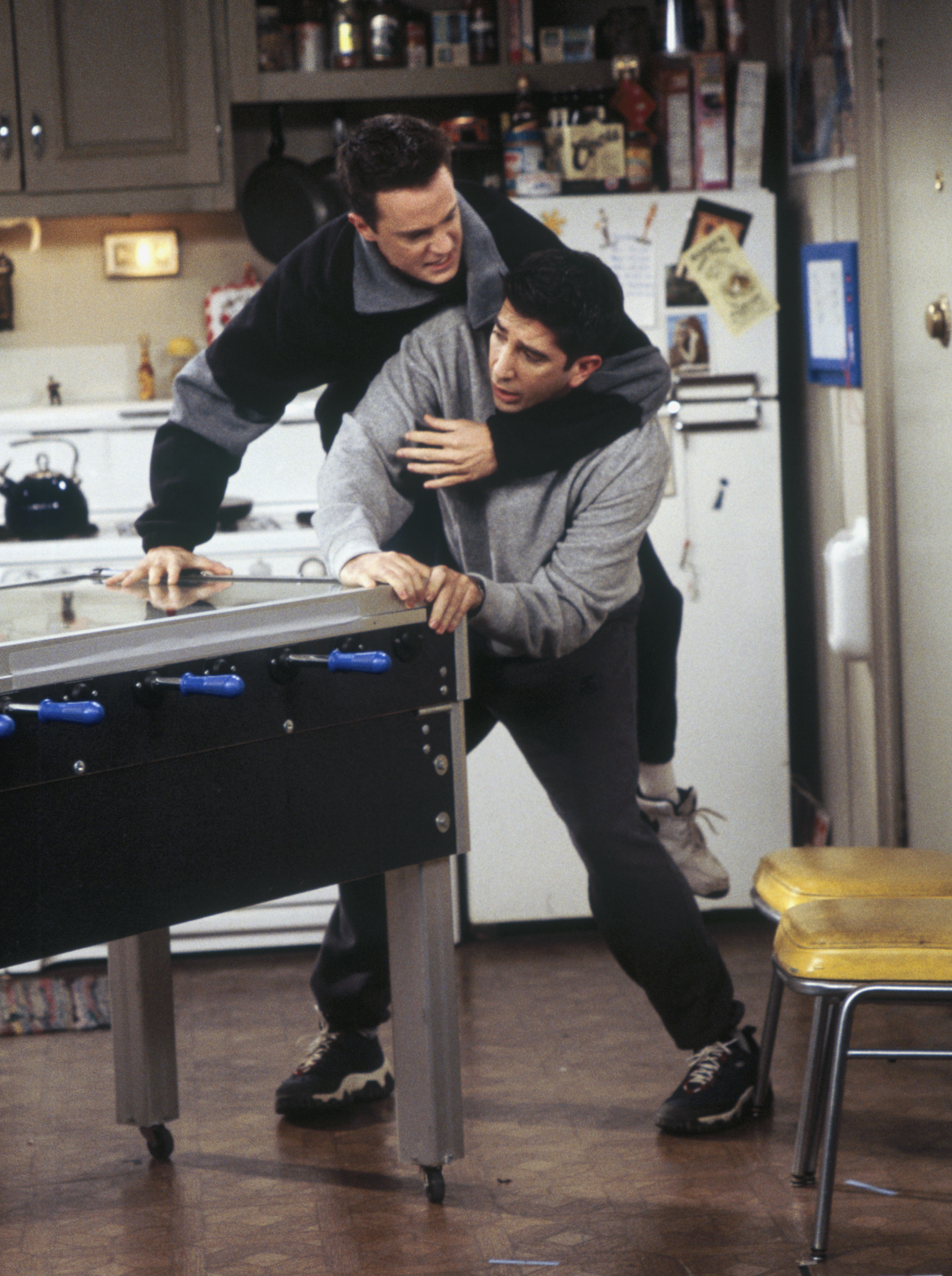 Matthew Perry and David Schwimmer in a playful wrestling pose by a foosball table on set