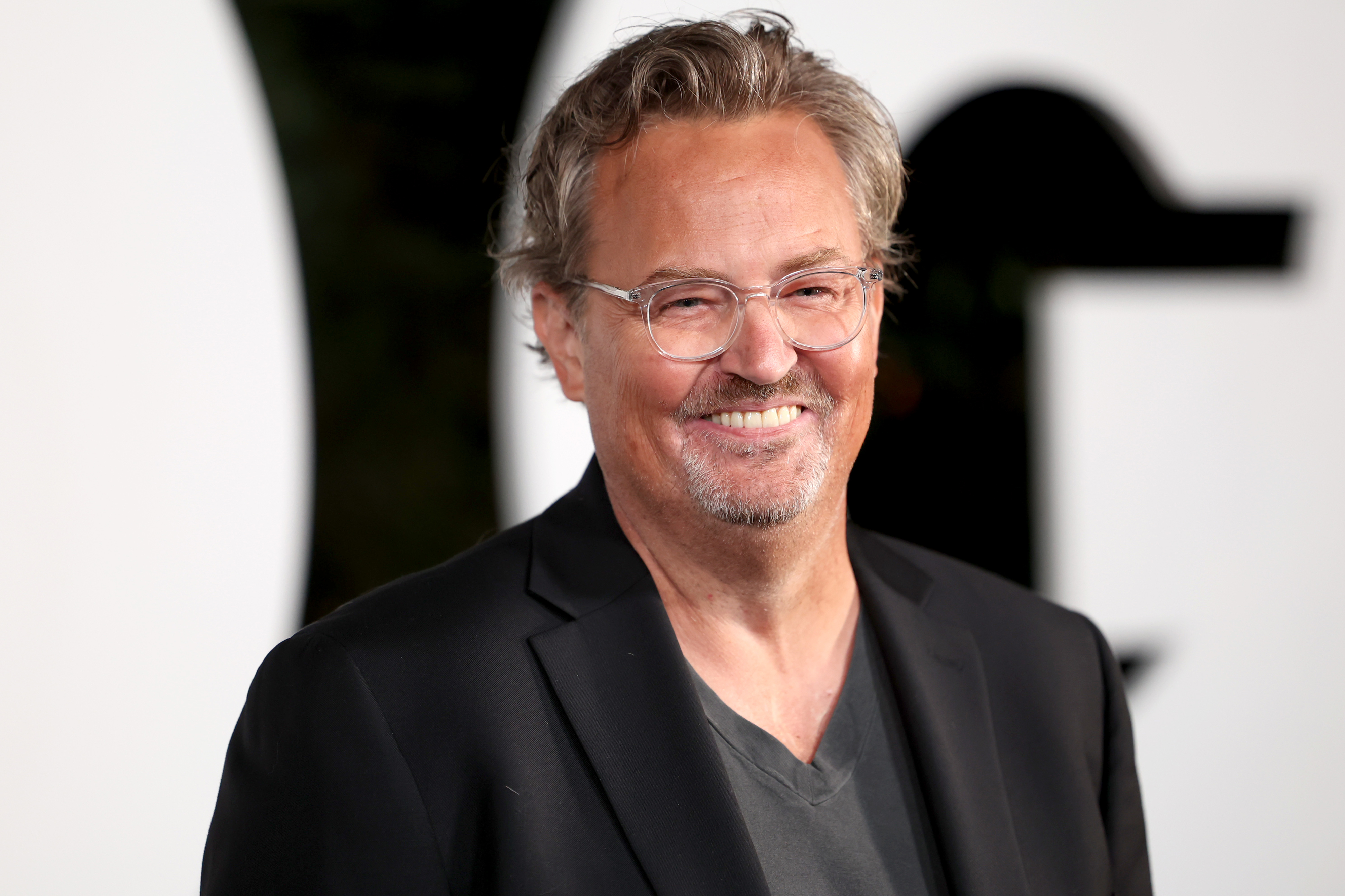 Matthew Perry smiles at the camera wearing a black blazer and a dark v-neck shirt at a celebrity event
