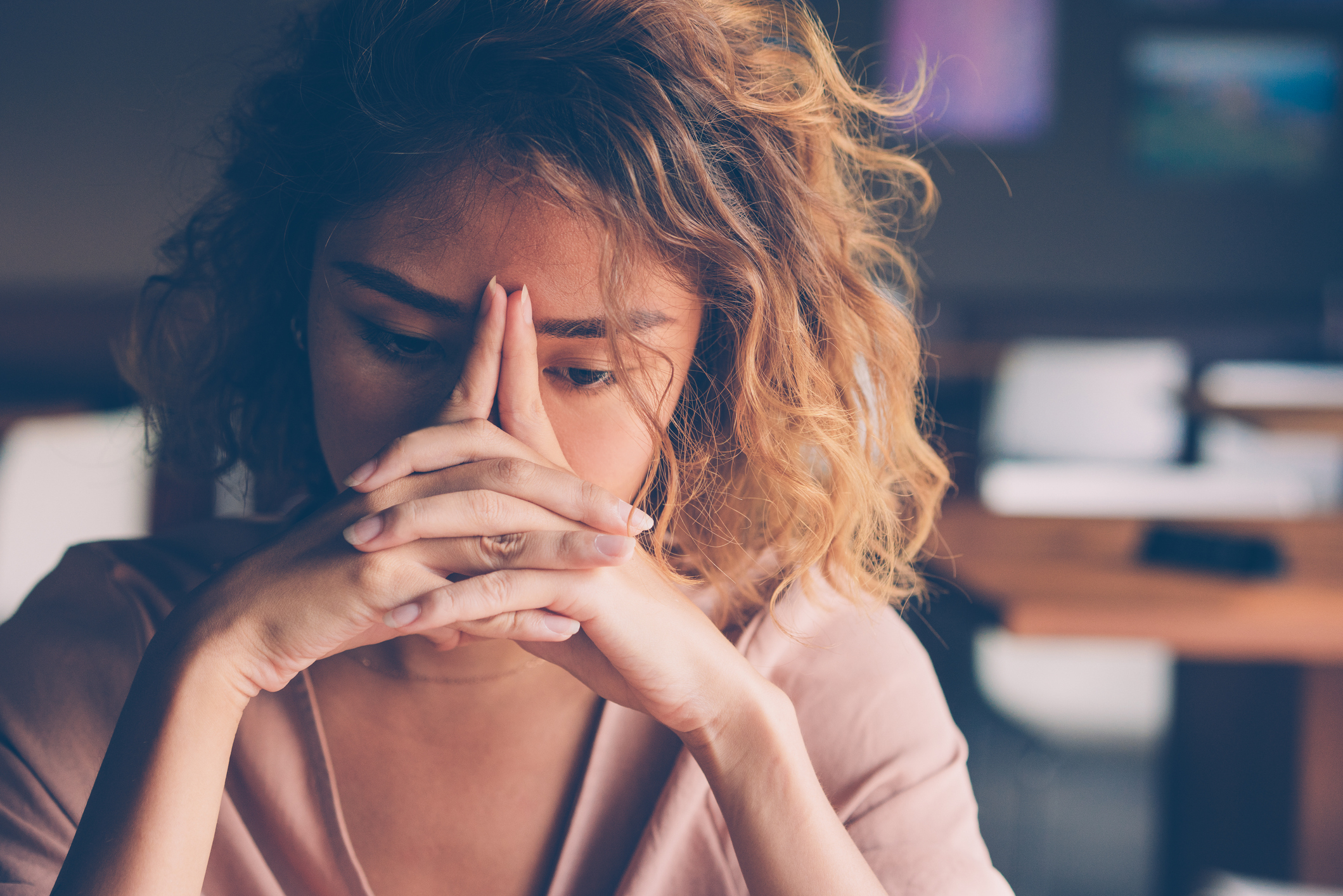 A woman sits at a table with her fingers pressed together in front of her face, appearing deep in thought or stressed