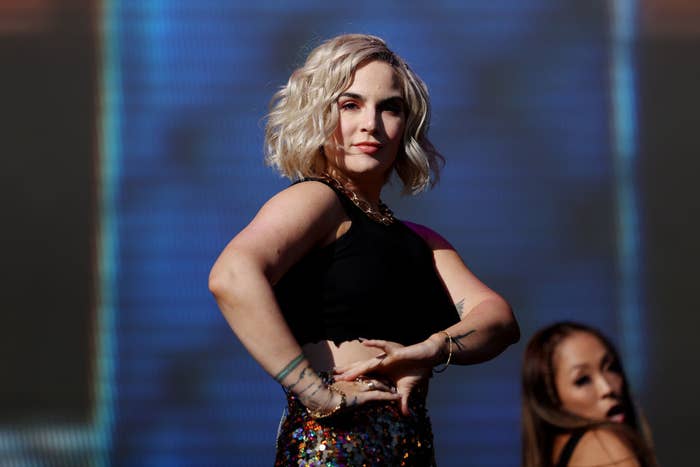 JoJo performs on stage, wearing a dark sleeveless top and a sequined skirt, with backup dancer visible behind her