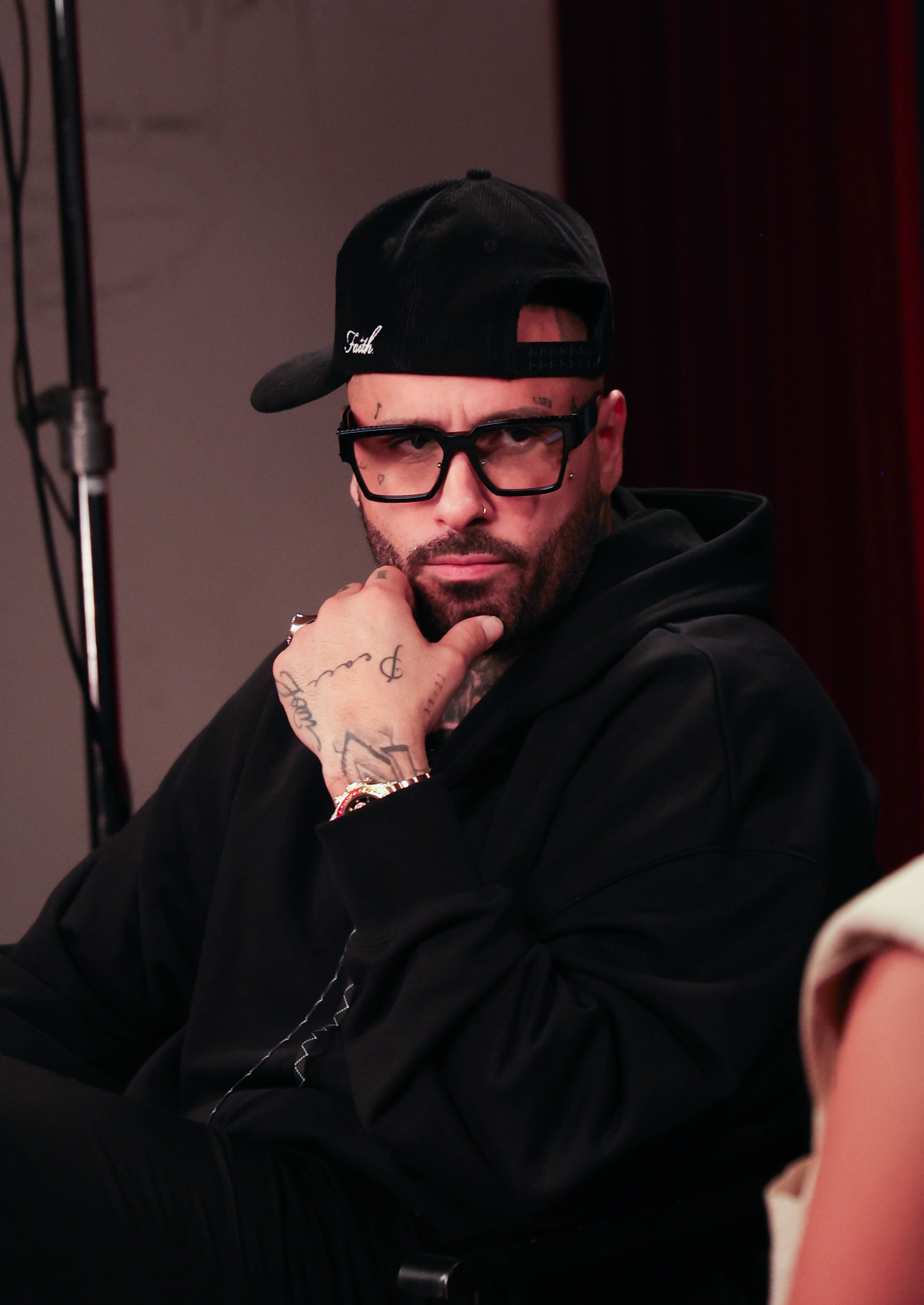 A person with glasses and a black cap worn backwards, dressed in a black hoodie, sitting and looking thoughtful with hand on chin