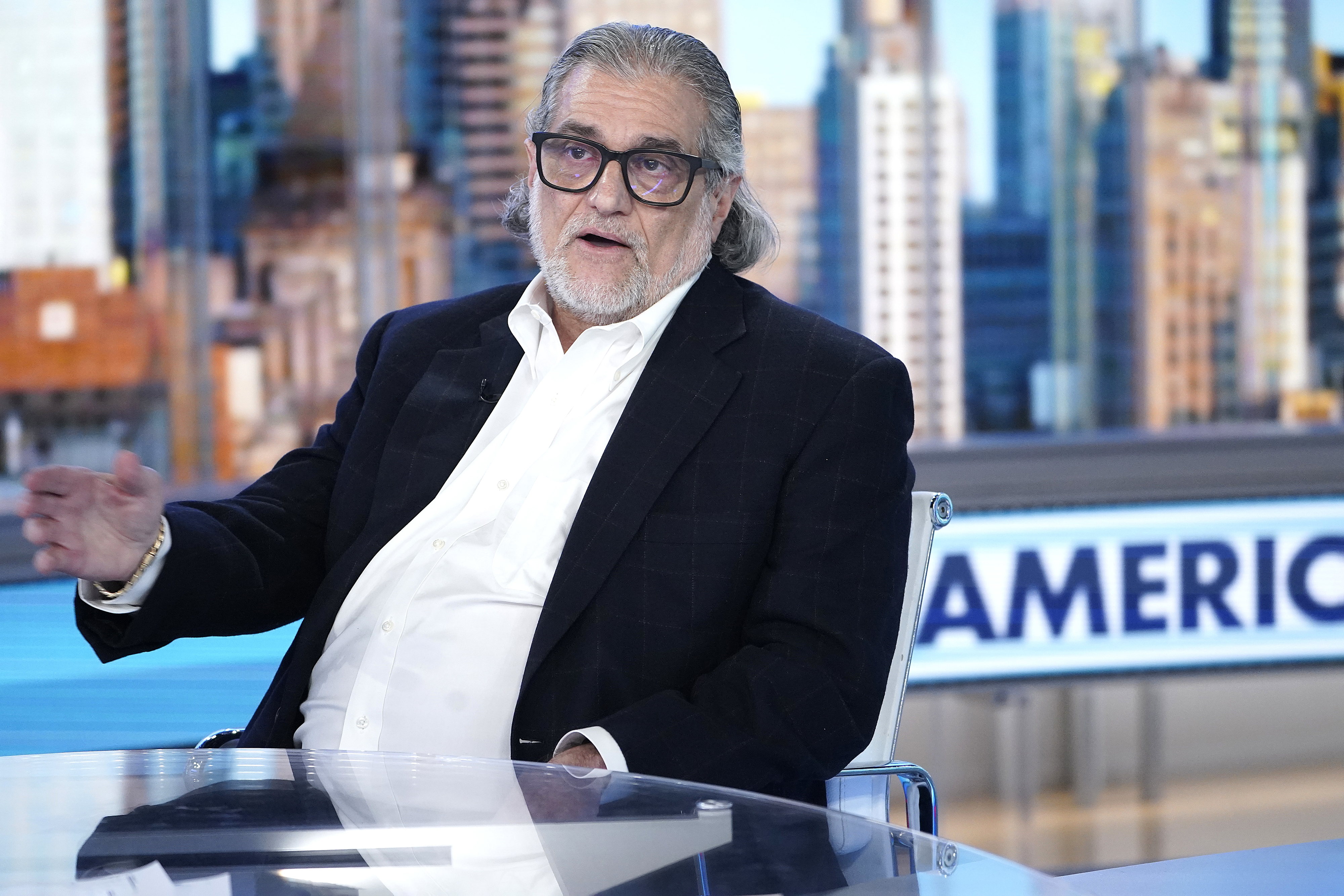 Man with gray hair and glasses, wearing a white shirt and dark blazer, speaks on a television set with a cityscape backdrop