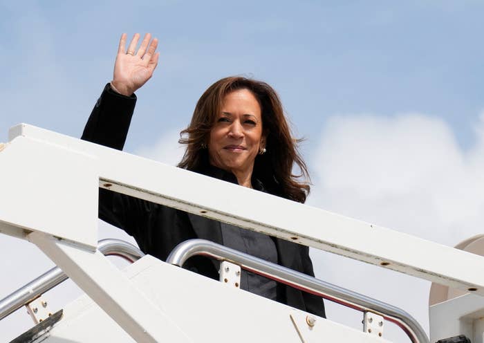 A woman wearing a dark blazer waves from the top of an airplane staircase