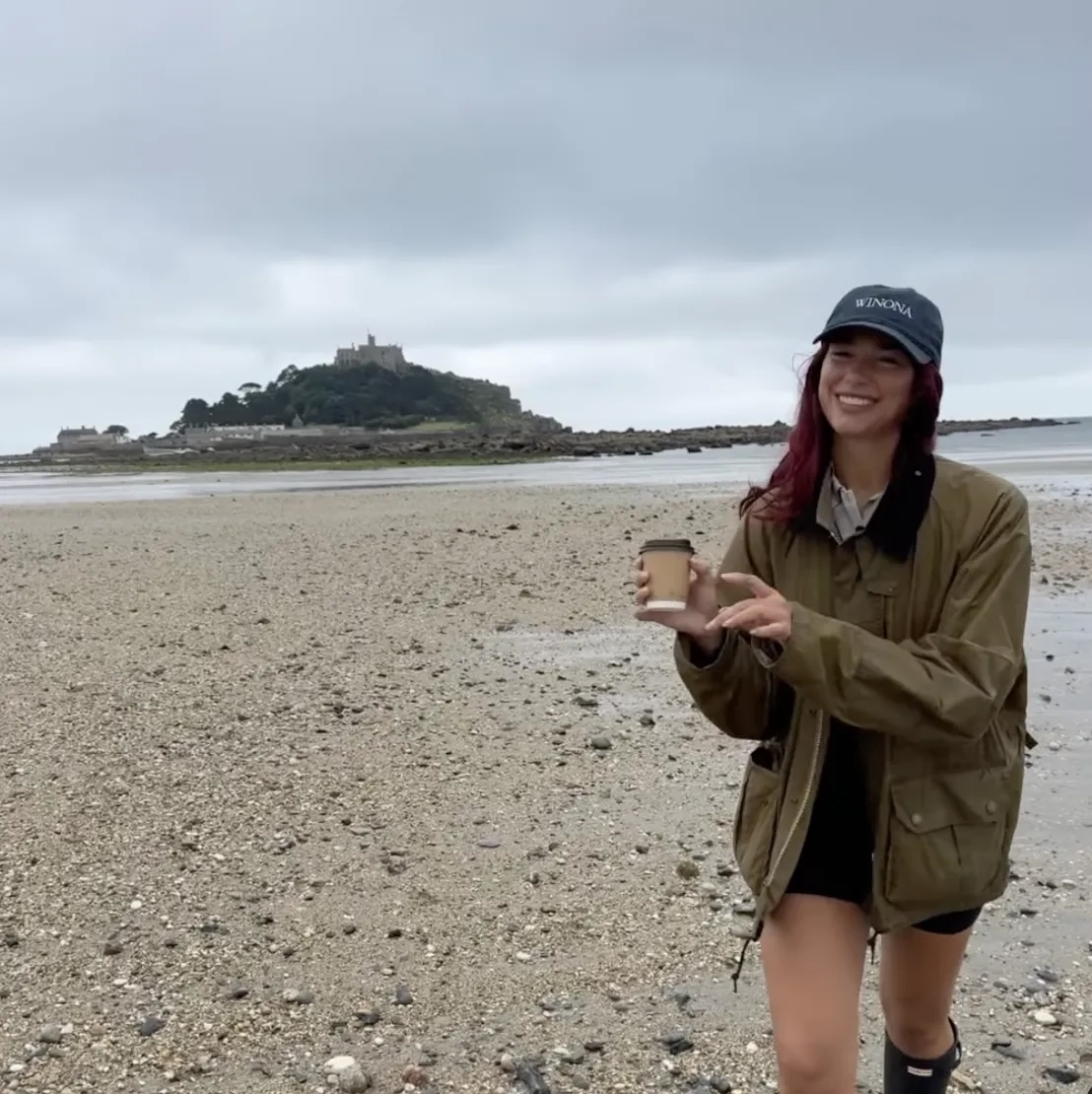 Dua Lipa smiles while holding a coffee cup on a rocky beach, wearing a green jacket, black shorts, and a dark hat. Island visible in the background