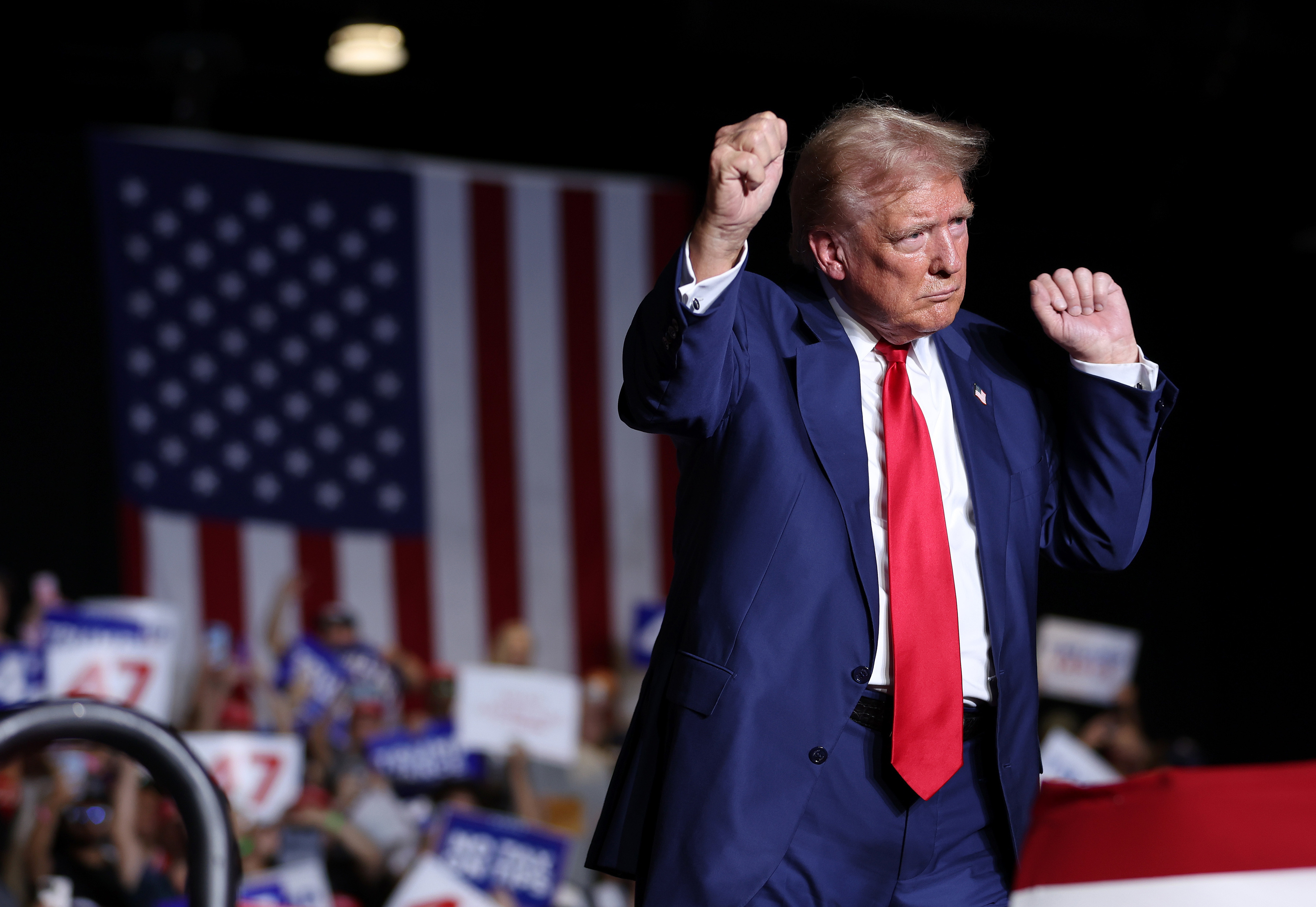 Donald Trump gestures with raised fists at a rally, standing in front of a crowd with an American flag in the background