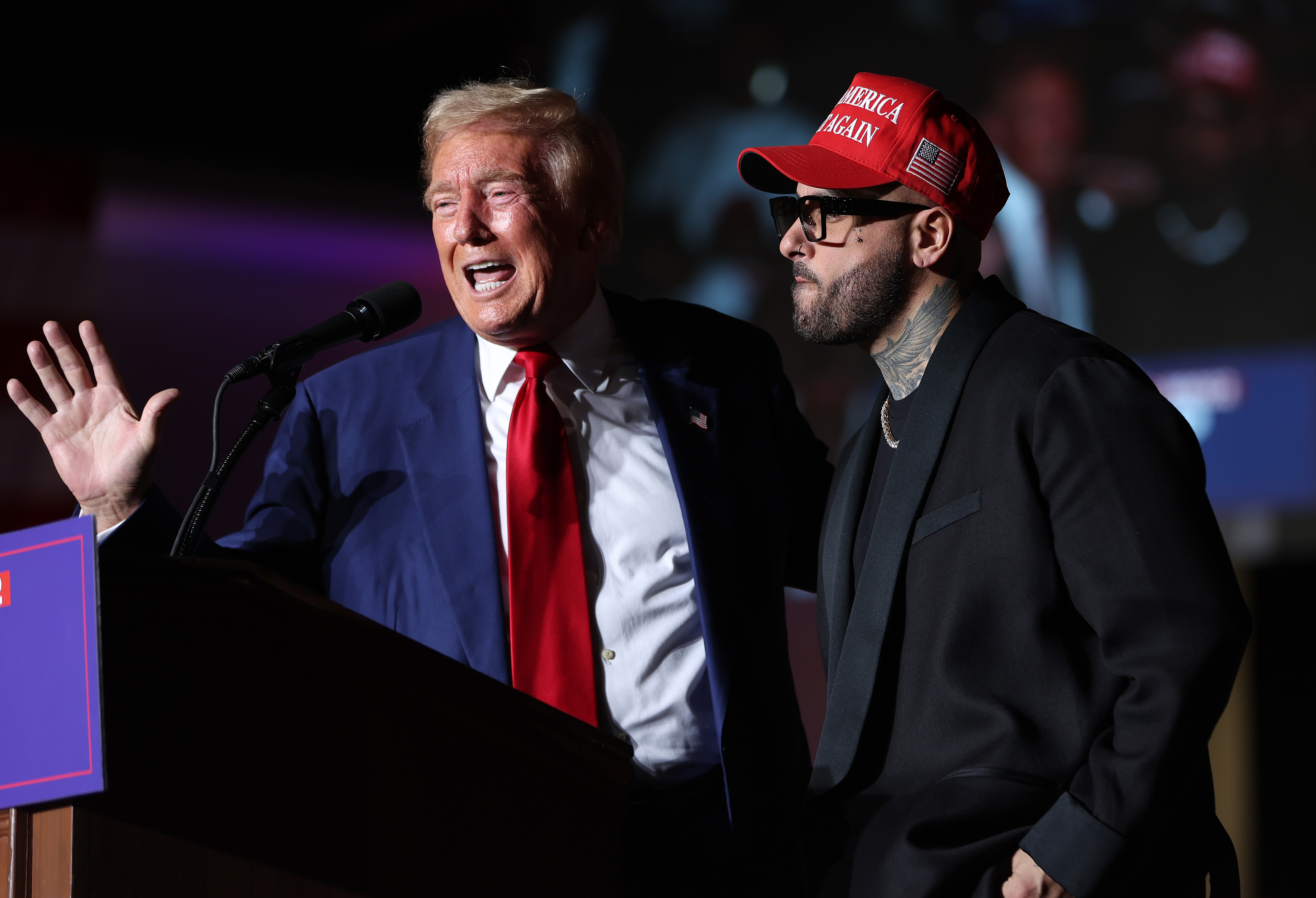 Donald Trump speaks at a podium as Nicky Jam, wearing a "Make America Great Again" hat, stands beside him