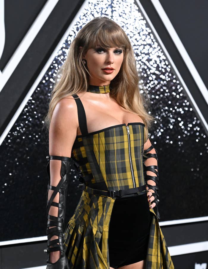 Taylor Swift in a plaid corset dress with black straps, leather gloves, and a choker on a black and white background at a public event