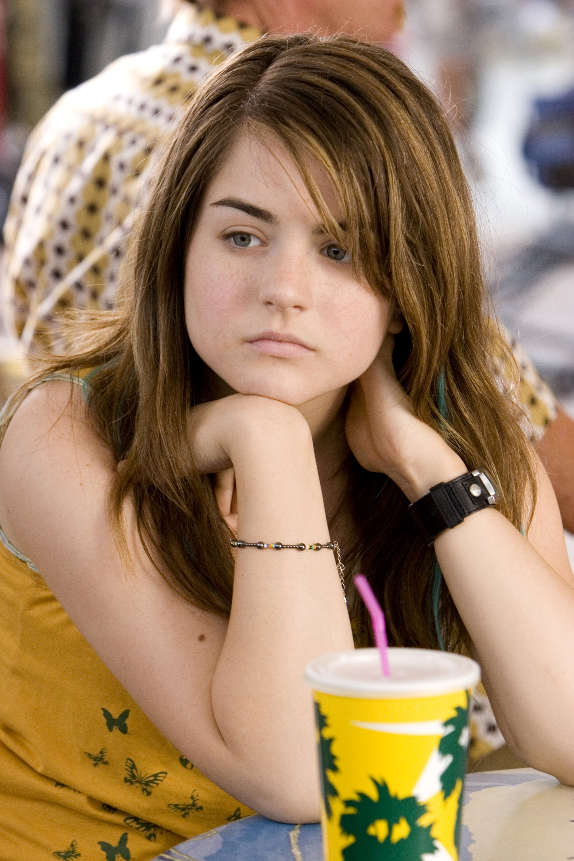JoJo sits at a table resting her chin on her hands, with a contemplative expression. A drink with a straw is in front of her