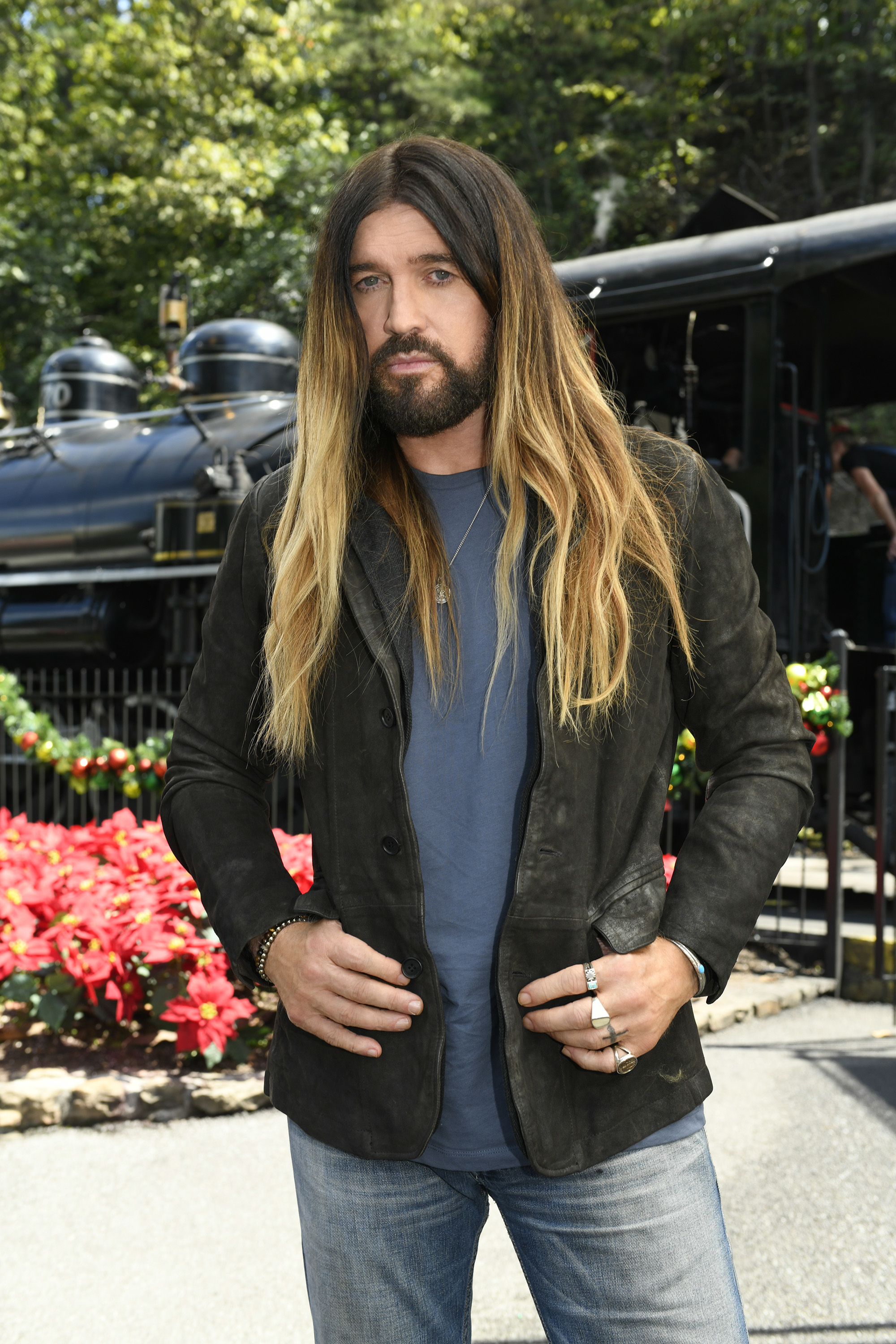 Billy Ray Cyrus poses outdoors in casual attire, wearing a blue shirt, leather jacket, and jeans, with a locomotive in the background