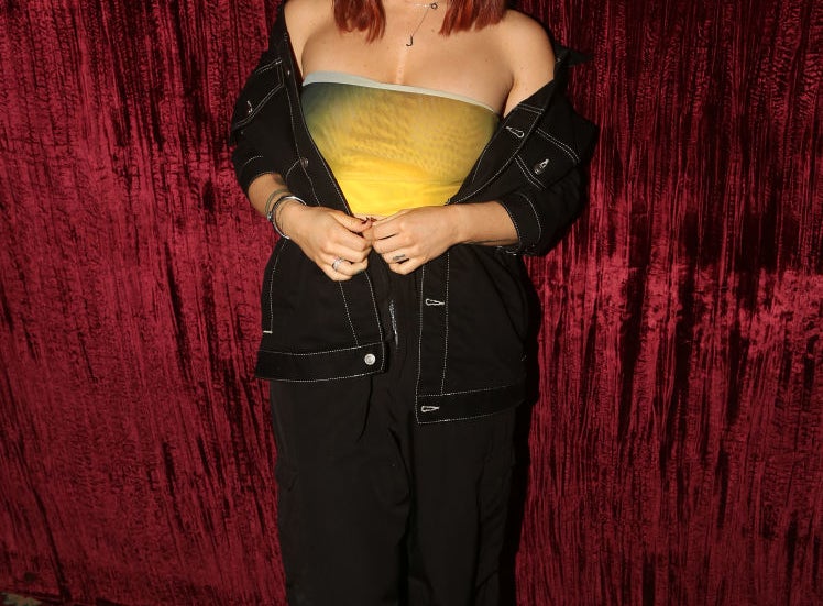 JoJo wears a strapless gradient top, dark jacket, and black wide-leg pants, posing in front of a textured backdrop