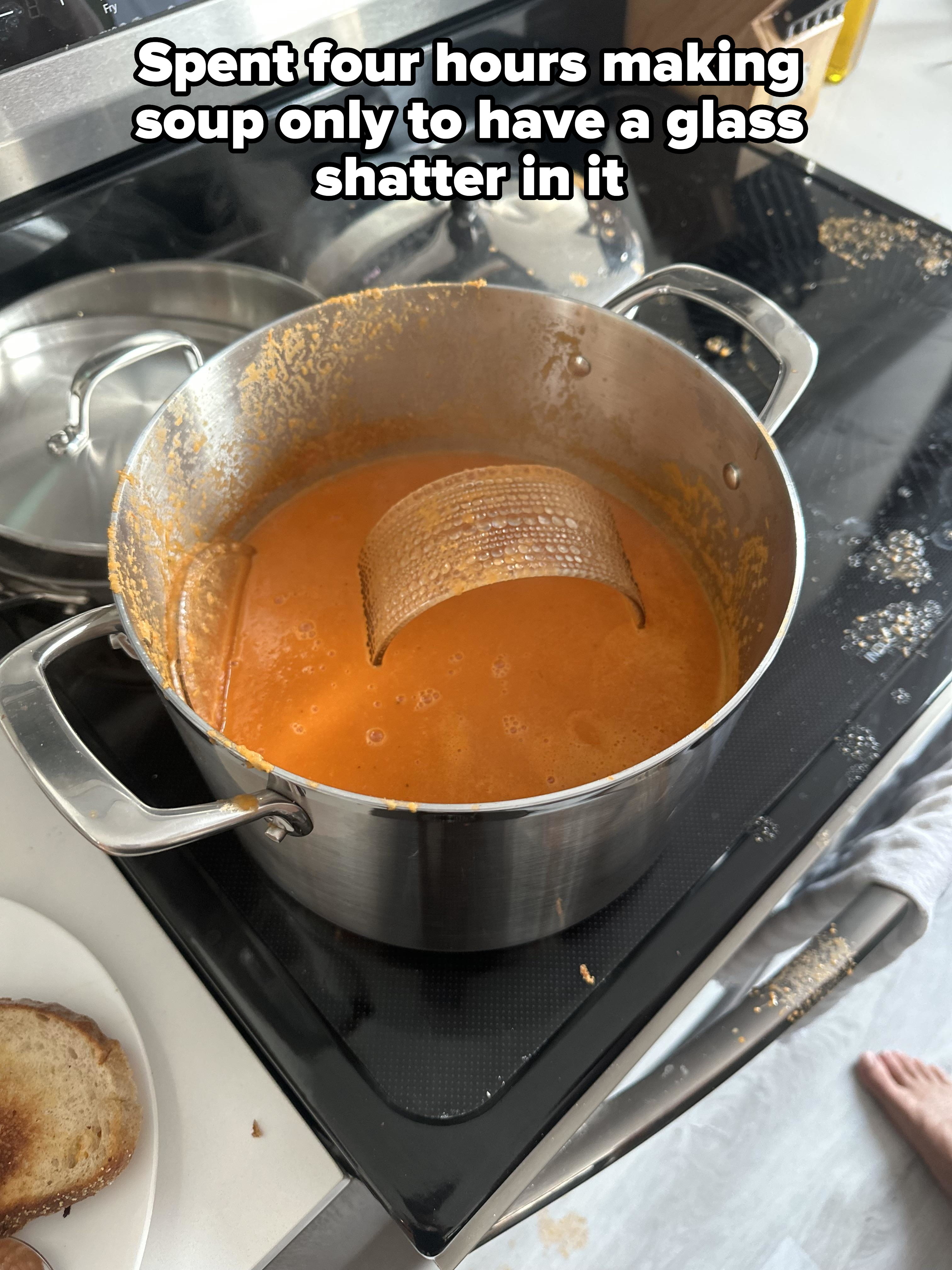 A large metal pot on a stove contains orange soup with part of a metal sieve submerged. Toast is on a plate next to the pot