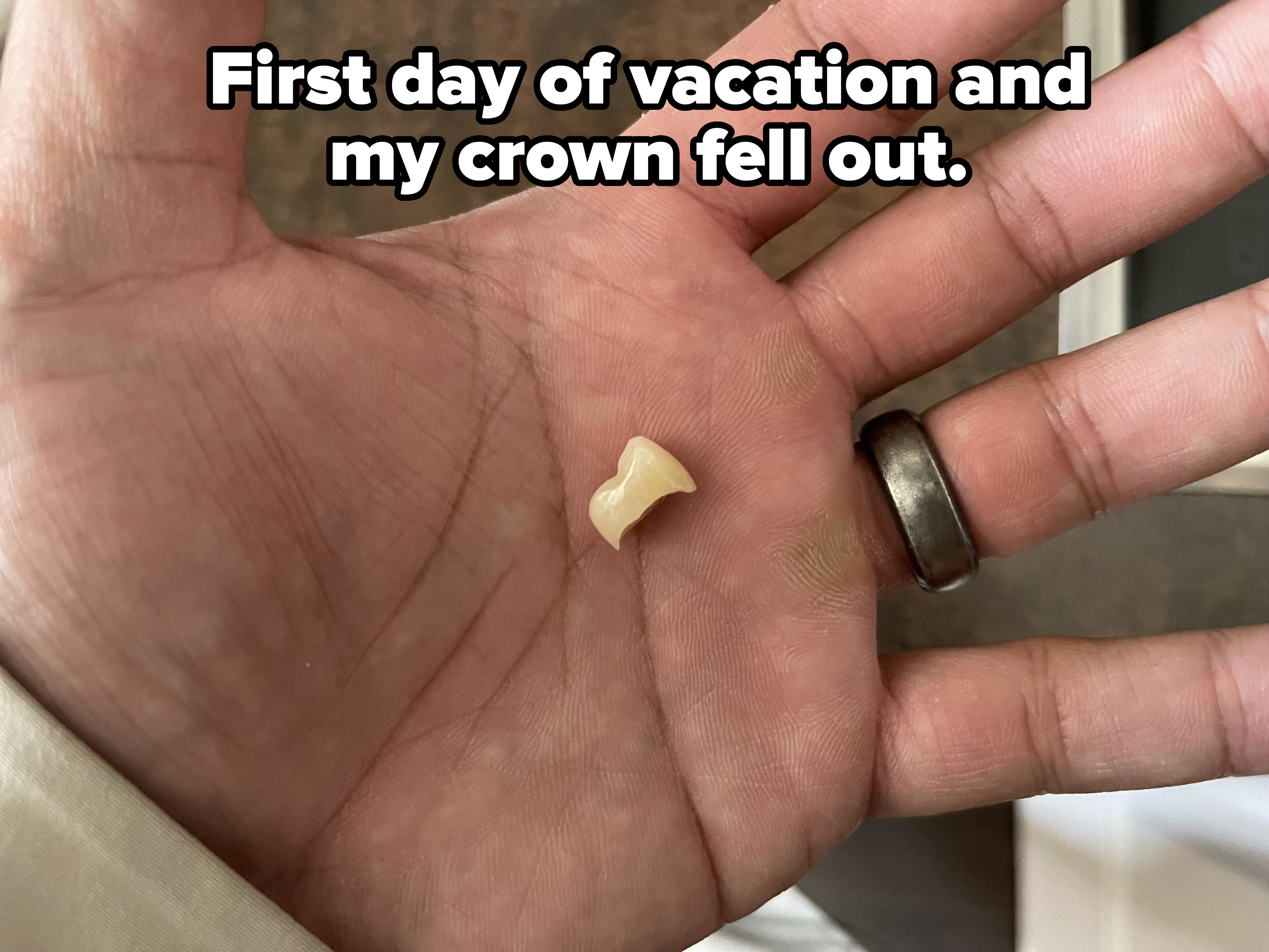 A hand holding a small, off-white pasta shell. The person is wearing a ring on their ring finger. The background is blurred