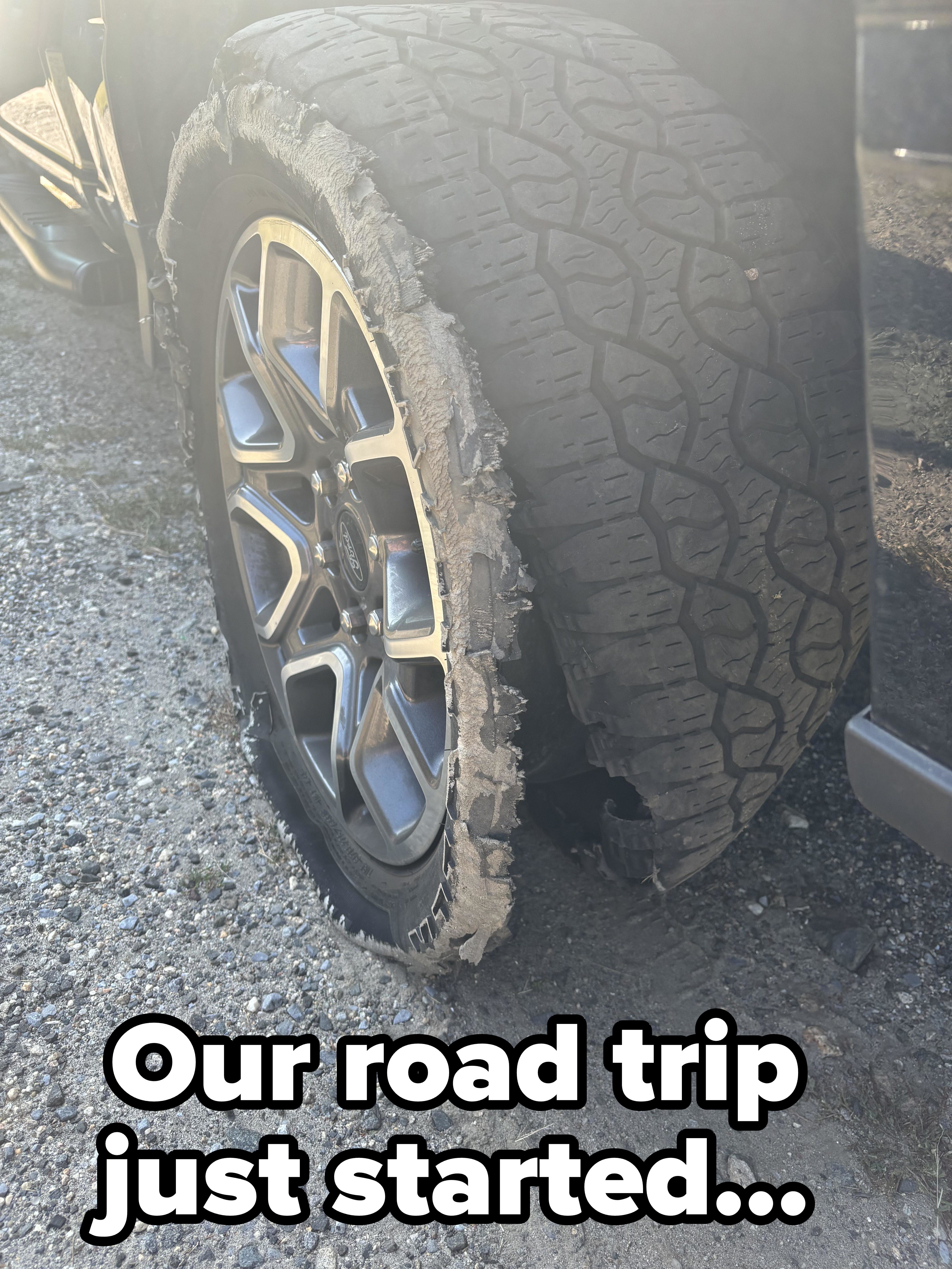 Close-up of a car's flat tire with a shredded outer edge, parked on a gravel surface. No people are visible