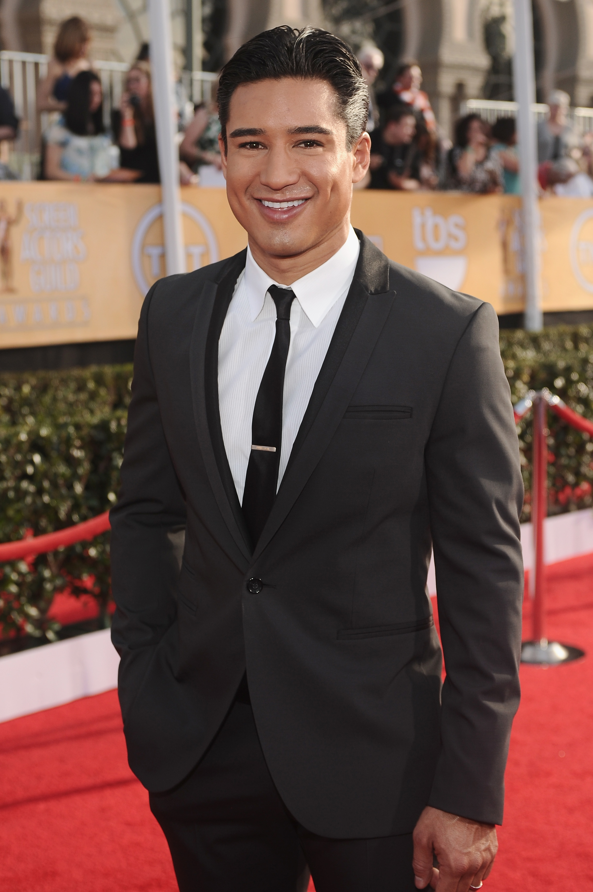Mario Lopez in a black suit and tie, posing on the red carpet at an event with a crowd and a gold backdrop behind him