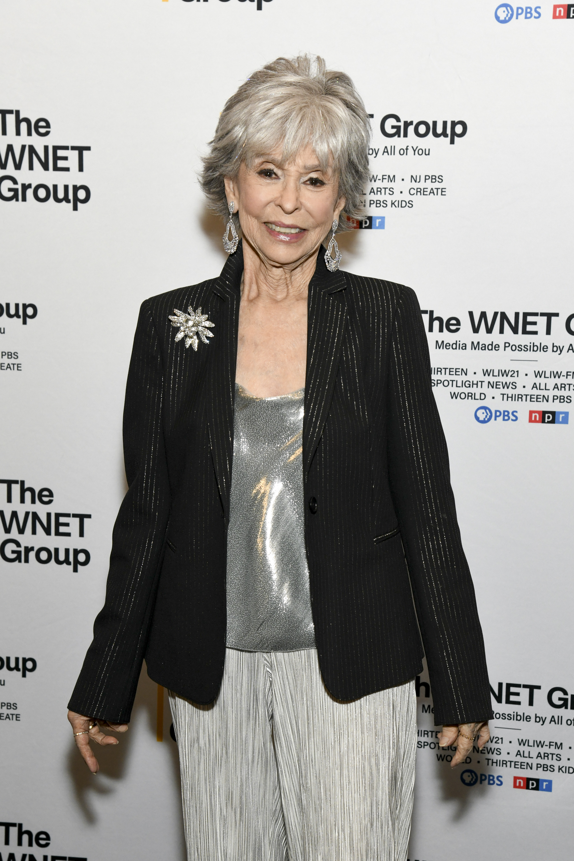 Rita Moreno on the red carpet, wearing a sparkling top, a black blazer with a brooch, and white pants, attending an event for The WNET Group