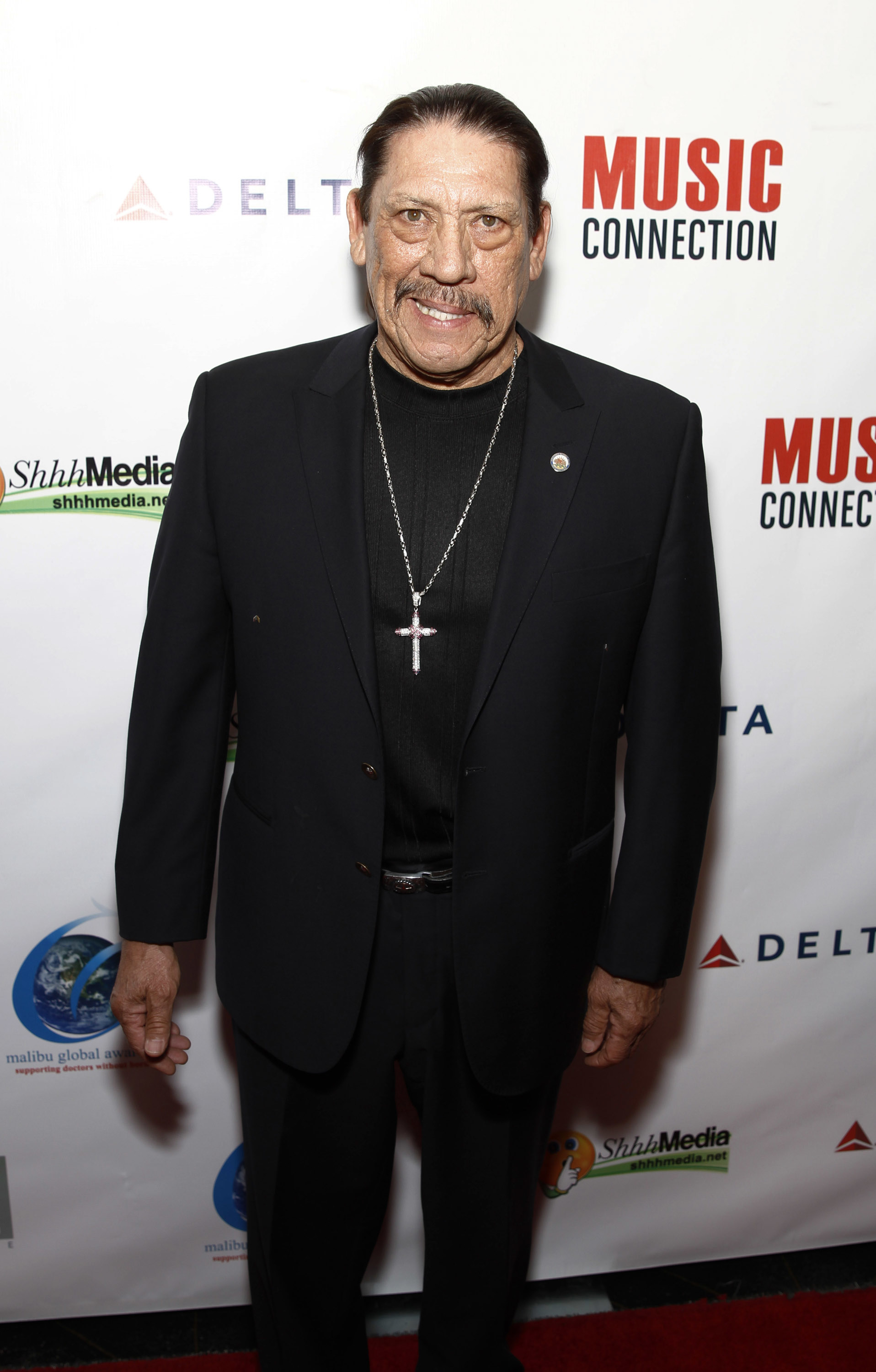 Danny Trejo on the red carpet at the Music Connection event, wearing a black suit and a cross necklace