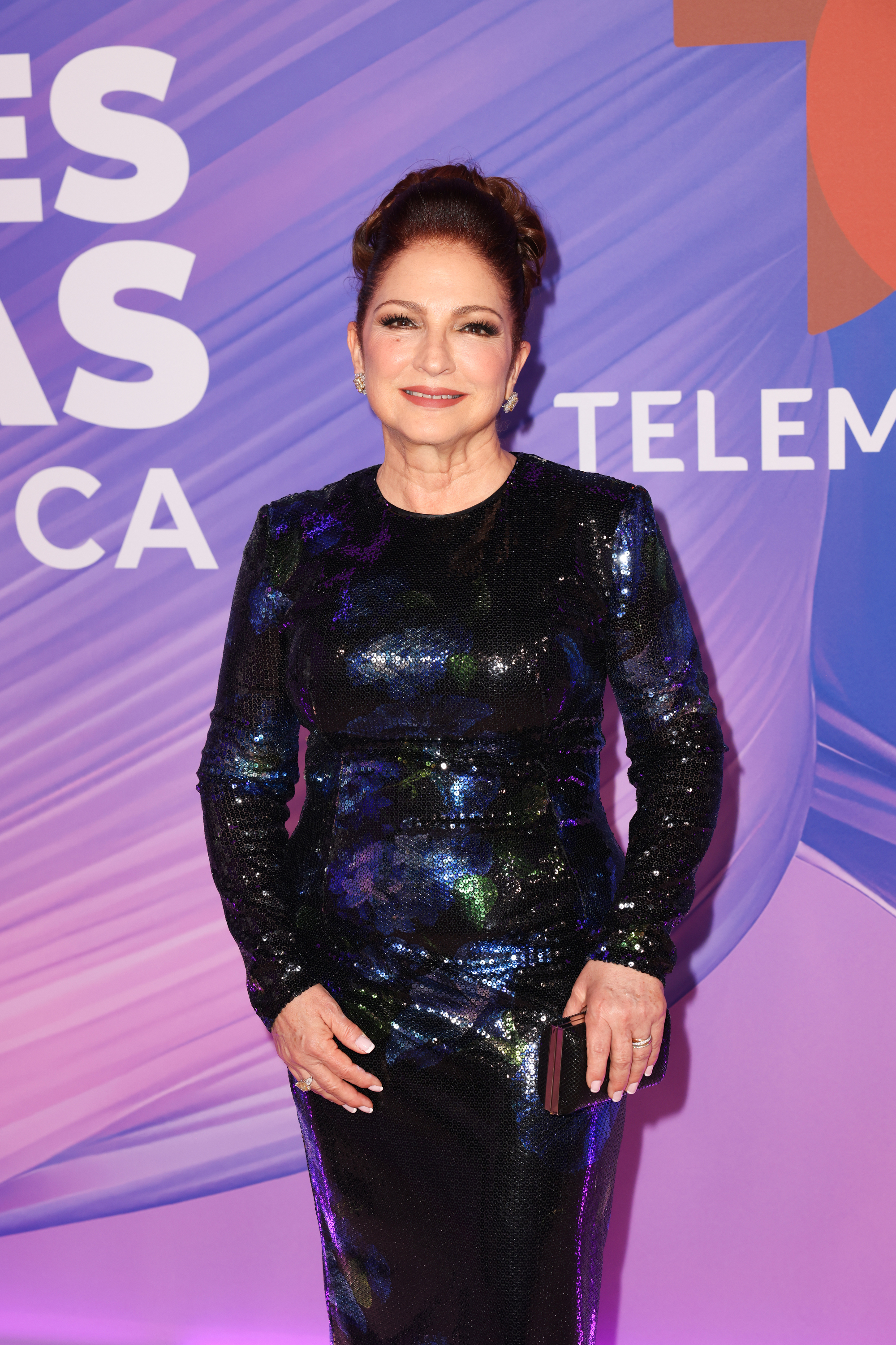 Gloria Estefan at a Telemundo event, wearing a long-sleeve, black sequined dress and holding a clutch. She is posing in front of a media backdrop