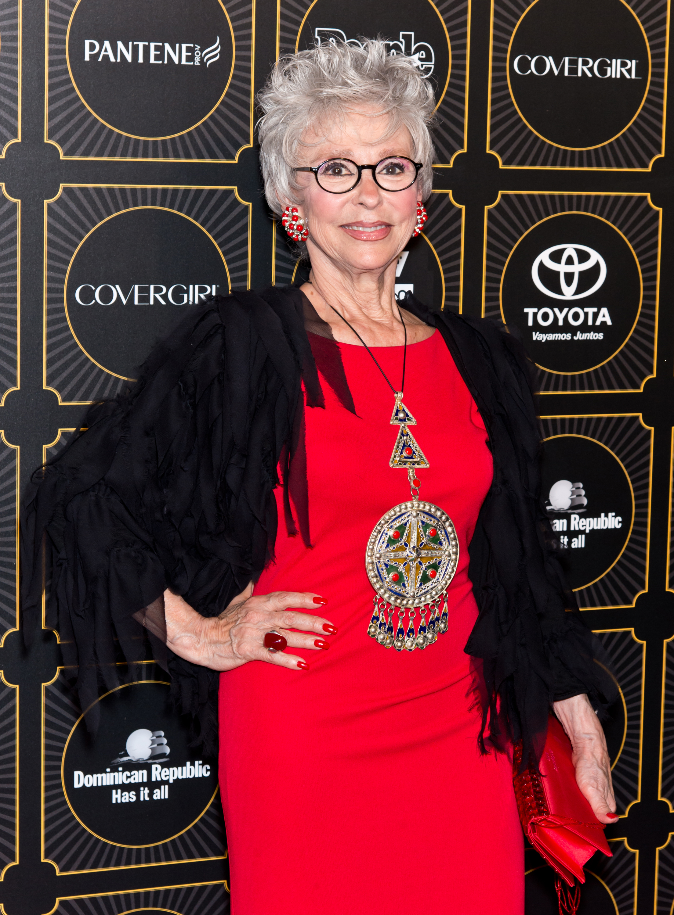 Rita Moreno on the red carpet in a red dress with a large pendant necklace at an event featuring sponsors like CoverGirl, Toyota, and Pantene