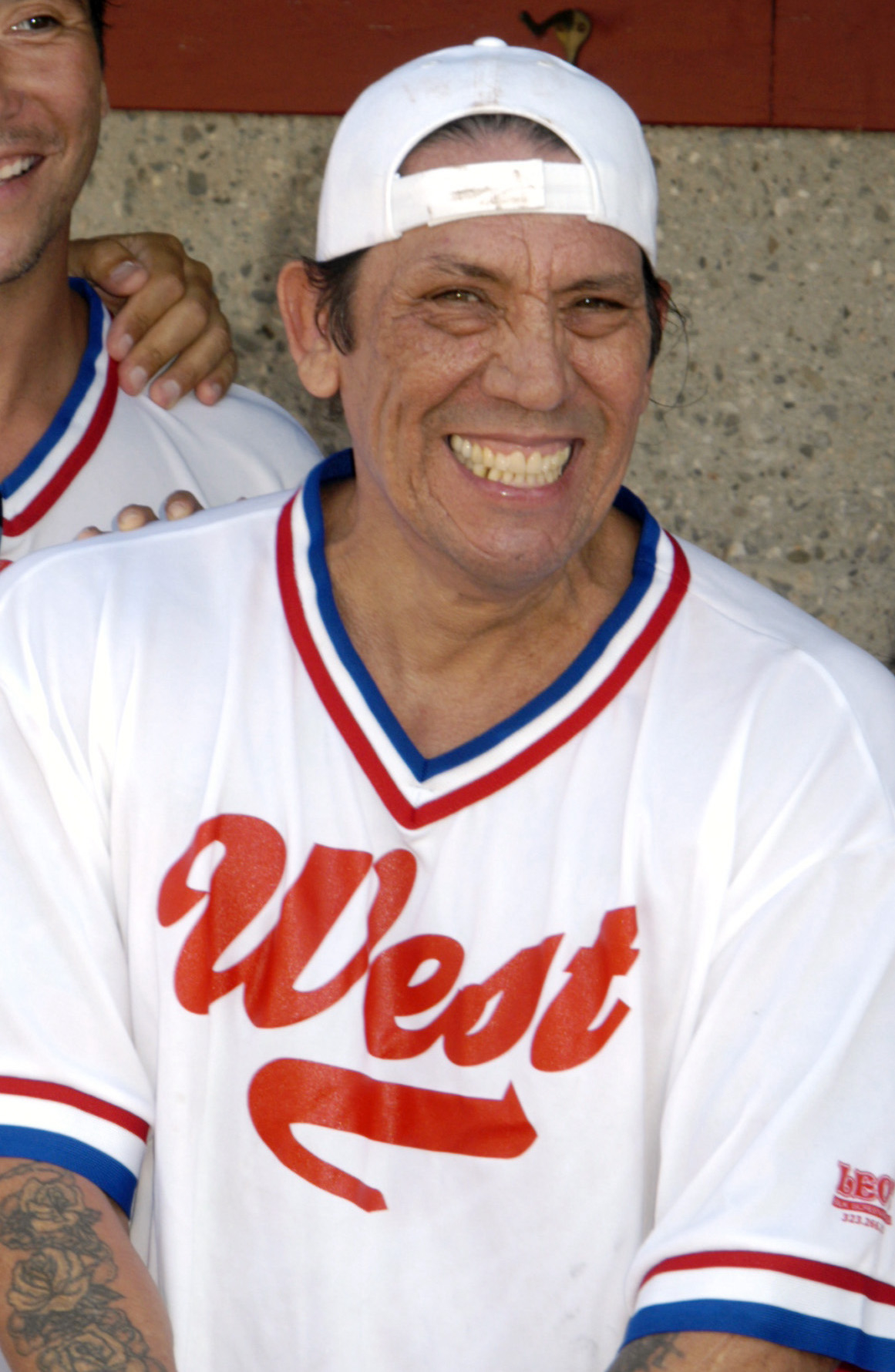 Danny Trejo smiling, wearing a white baseball jersey with "West" on it and a backward white cap