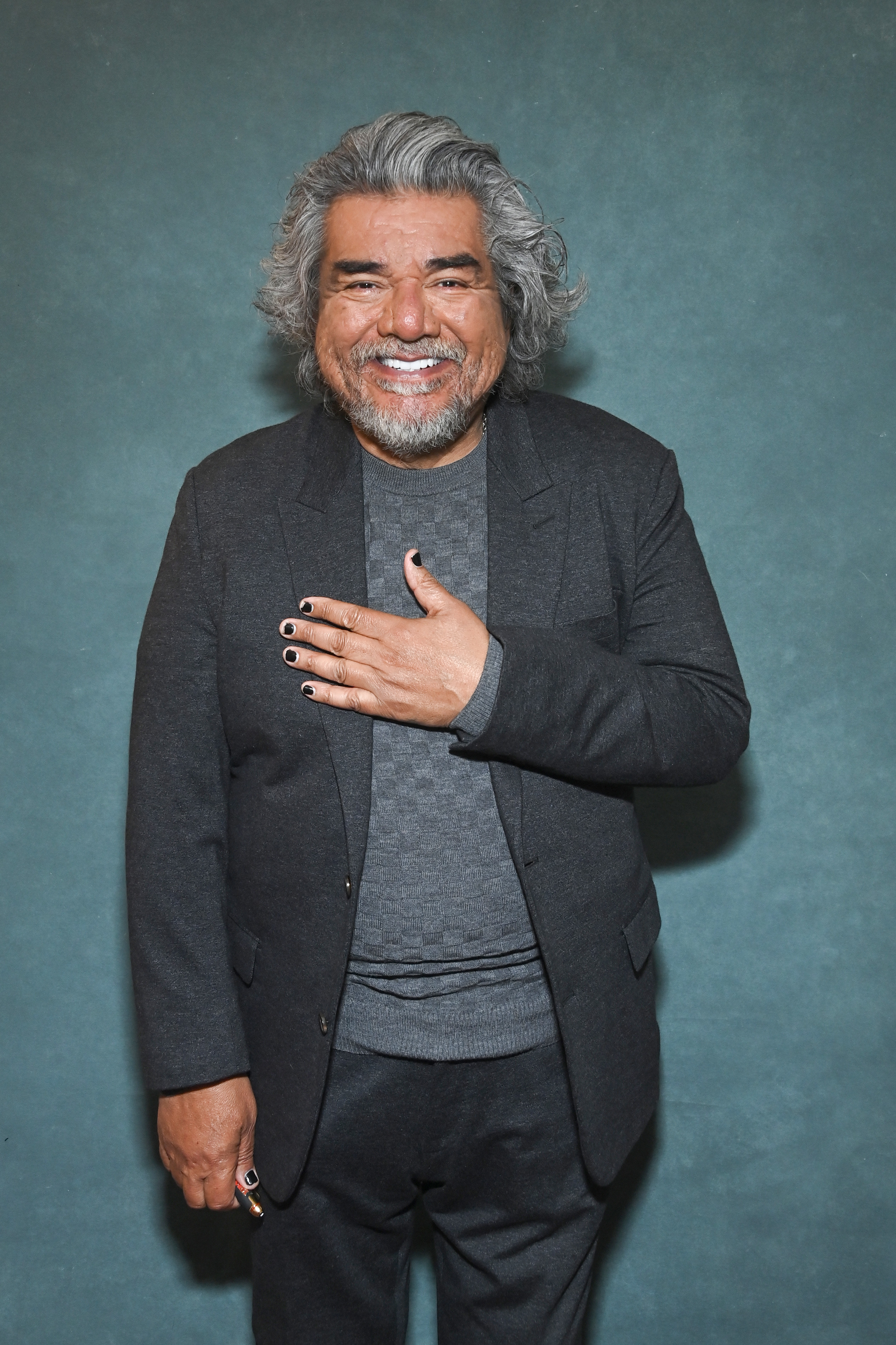 George Lopez smiles, wearing a dark suit with a gray shirt, posing with one hand on his chest, nails painted black